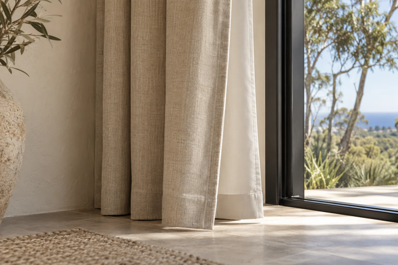 Oatmeal curtain hem and pale lining meeting the floor beside a black-framed window.