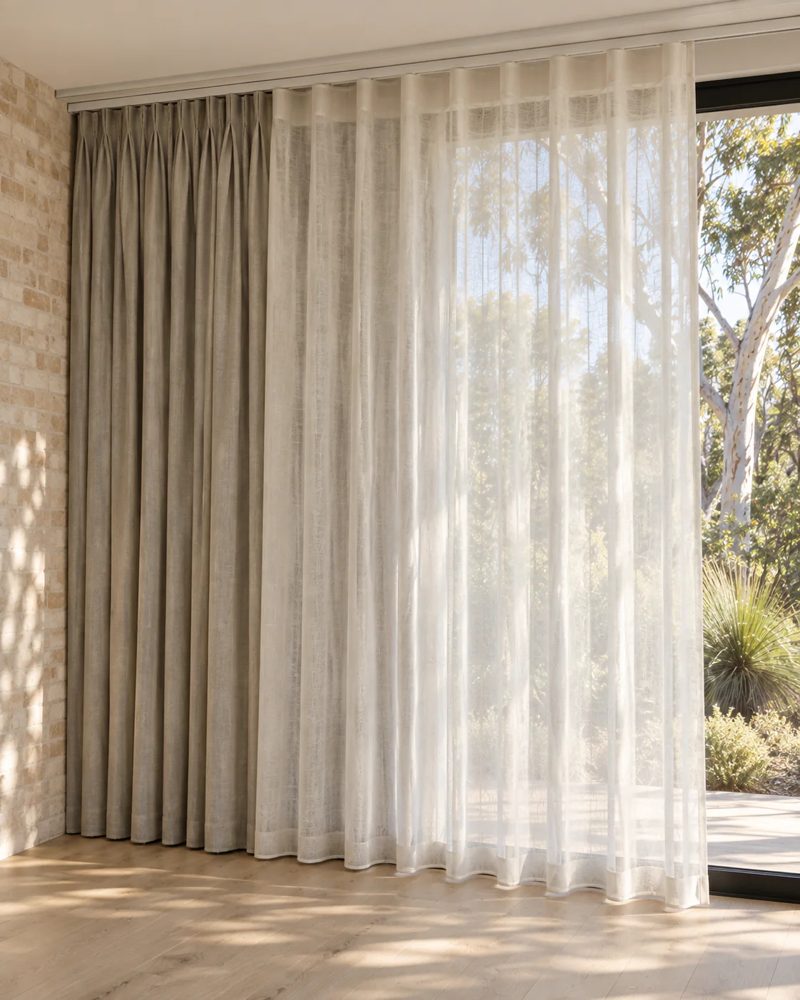 Room-side ivory sheers overlapping rear stone curtains beside a brick wall and black-framed window.