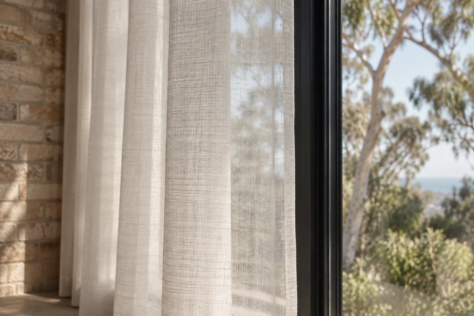 Daylight passing through the open weave of an ivory sheer beside a black window frame.