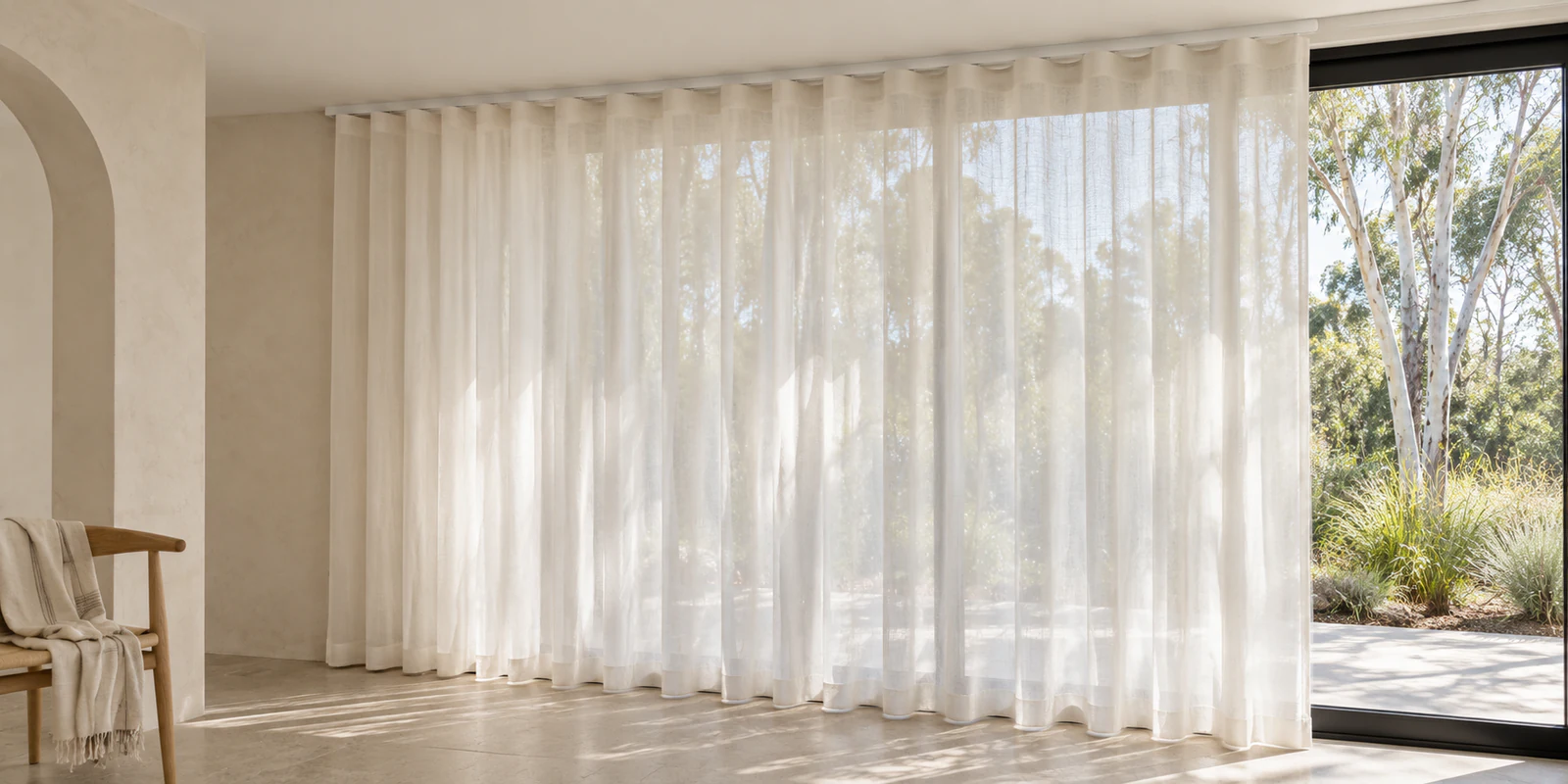 Full-height ivory sheer curtains hanging in soft waves below a visible track, with native greenery softly visible through the fabric.