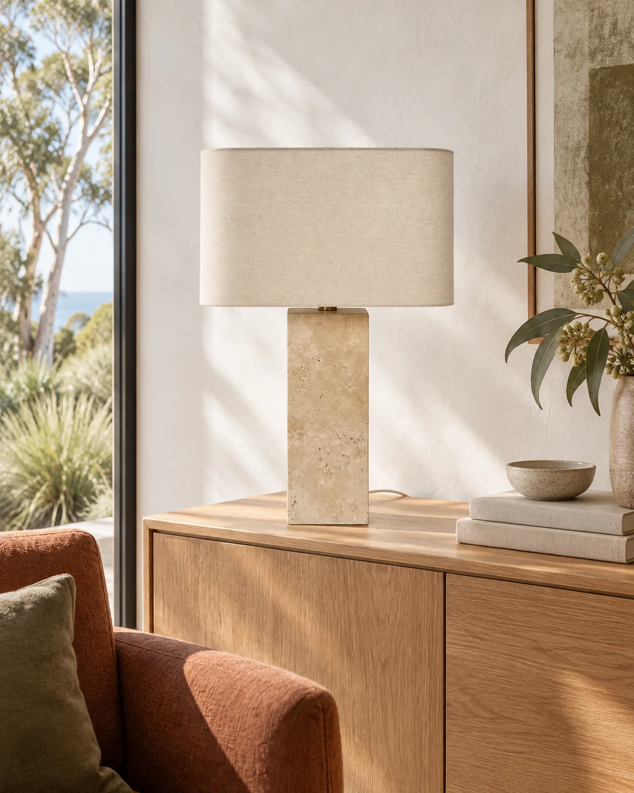 Travertine table lamp with a linen shade on an oak sideboard beside a rust armchair