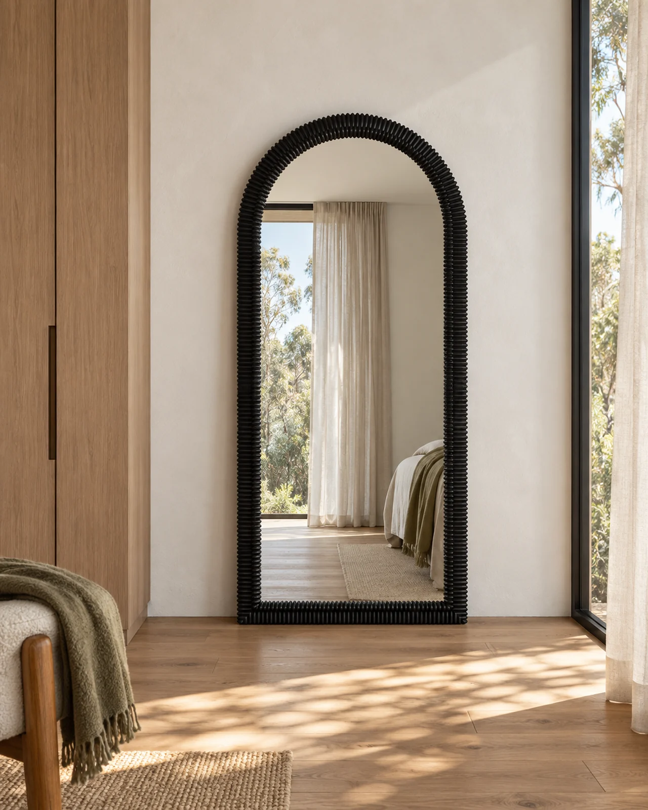 Black ribbed arched mirror beside an oak wardrobe, reflecting a sunlit bedroom